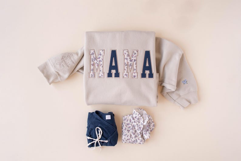 A sand colored sweatshirt with the word MAMA embroidered with baby clothes as a keepsake sweatshirt and baby names on the sleeve. The image also includes the text customize with your baby clothes
