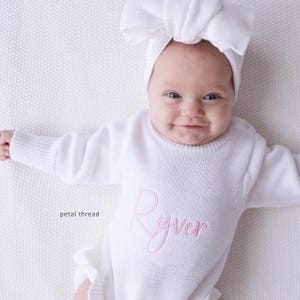 Pu&ograve; includere: Una tutina in maglia bianca a maniche lunghe con il nome Ryver ricamato in corsivo rosa sul petto, abbinata a una fascia bianca coordinata con un grande fiocco. Il testo petal thread &egrave; visibile sul lato sinistro dell'immagine.