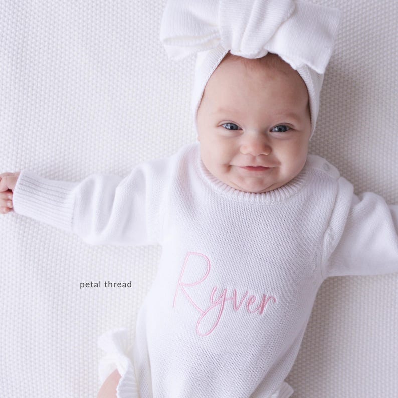 Pu&ograve; includere: Una tutina in maglia bianca a maniche lunghe con il nome Ryver ricamato in corsivo rosa sul petto, abbinata a una fascia bianca coordinata con un grande fiocco. Il testo petal thread &egrave; visibile sul lato sinistro dell'immagine.