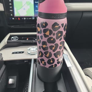 May include: A pink and black leopard print water bottle with a black lid and yellow accents. The bottle is in a car's cup holder. The brand name "Owala" is printed on the bottle.