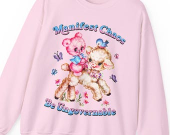 May include: Pink sweatshirt with the text "Manifest Chaos Be Ungovernable." The graphic features a pink teddy bear riding a lamb with a blue bird on its head. Butterflies and flowers surround the characters.