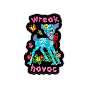 May include: Embroidered patch featuring a turquoise deer with a red bow, yellow flower, and butterflies. The patch has the words "wreak havoc" in pink lettering on a black background. The deer is surrounded by colorful flowers.