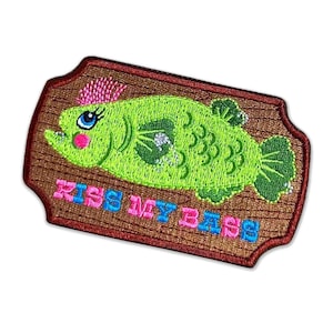 May include: An embroidered patch featuring a lime green fish with pink hair and blue eyes on a brown wood-grain background. The text KISS MY BASS is stitched in alternating pink and blue letters below the fish. The patch has a dark red border.