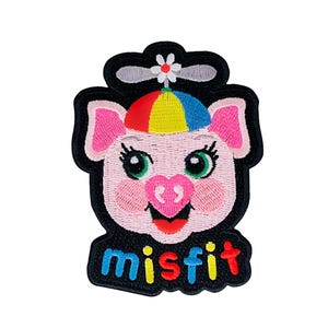 May include: Embroidered patch featuring a cartoon pig with a colorful hat and the word "misfit." The pig has pink ears, a heart-shaped nose, and green eyes. The hat is rainbow-colored, and the patch has a black border.