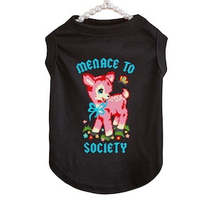 May include: Black pet shirt with a graphic of a pink deer with a blue bow, a butterfly, and the words "MENACE TO SOCIETY" in turquoise. The shirt is sleeveless and hangs from a pearl beaded hanger.