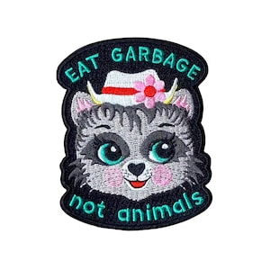 May include: Embroidered patch featuring a cartoon raccoon wearing a hat with a pink flower. The patch has the text "EAT GARBAGE not animals" in teal lettering. The raccoon has teal eyes and pink cheeks, set against a black background.