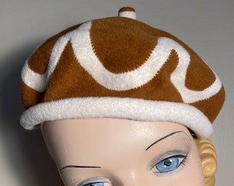 Peut inclure: Un béret marron avec un motif blanc ondulé. Le béret est porté sur un mannequin à la peau claire, aux yeux bleus et au rouge à lèvres rouge.