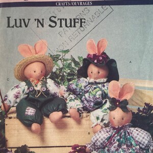 Peut inclure: Trois poupées de lapin en tissu faites à la main, habillées de tenues différentes. Les poupées sont assises sur une caisse en bois avec des plantes en pot et des carottes devant elles. Le modèle s'appelle "Luv 'N Stuff" et est de Butterick 6615.