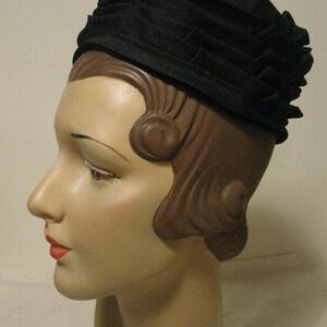 Puede incluir: Un sombrero de tela negro con un dise&ntilde;o plisado. El sombrero se lleva en un maniqu&iacute; con el pelo marr&oacute;n peinado en un recogido vintage.