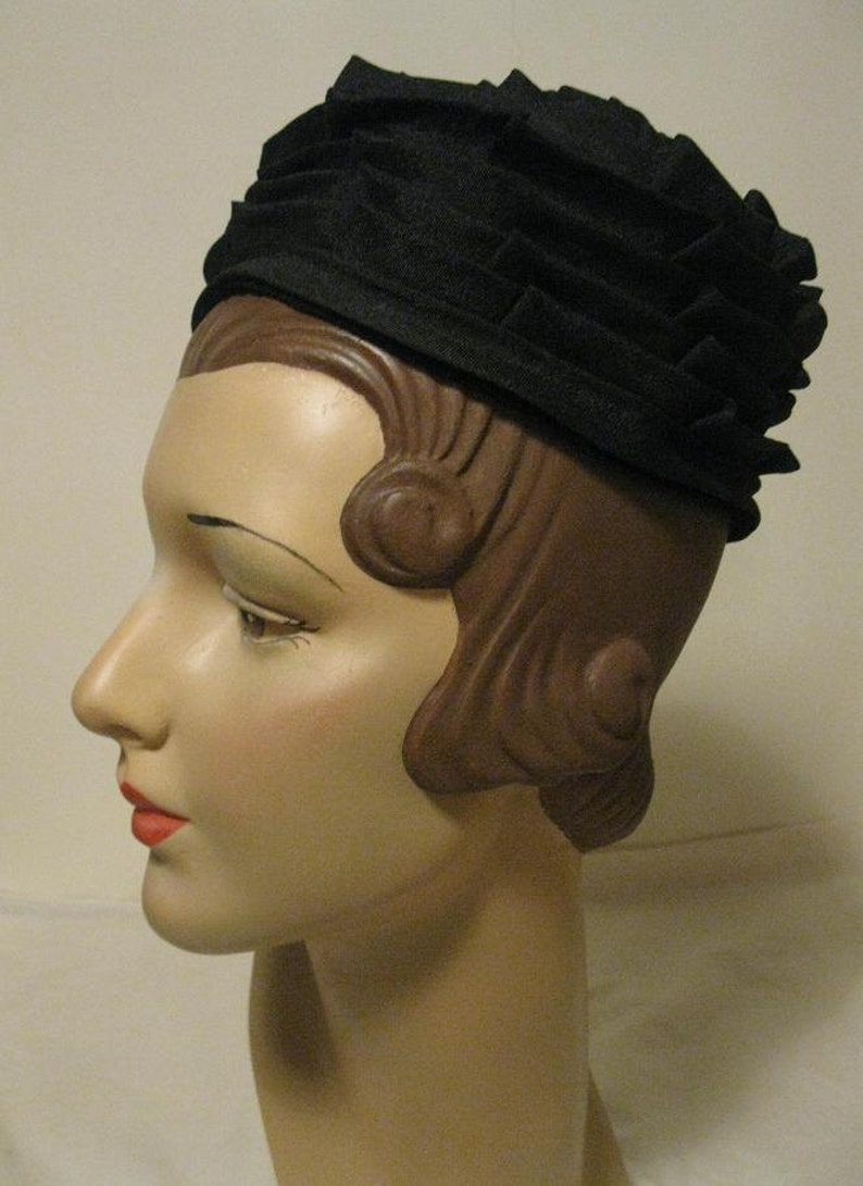Puede incluir: Un sombrero de tela negro con un dise&ntilde;o plisado. El sombrero se lleva en un maniqu&iacute; con el pelo marr&oacute;n peinado en un recogido vintage.