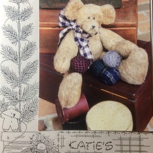 Peut inclure: Un ours en peluche beige avec un ruban à carreaux bleus et blancs autour du cou tient trois balles à carreaux. L'ours est assis sur une boîte en bois avec le texte "KATIE'S HOMESPUN STITCHES" sur le devant.