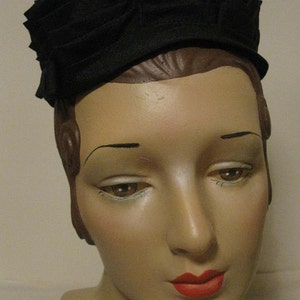 Puede incluir: Un sombrero de tela negro con un dise&ntilde;o plisado. El sombrero se lleva en un maniqu&iacute; con una expresi&oacute;n neutral y l&aacute;piz labial rojo.