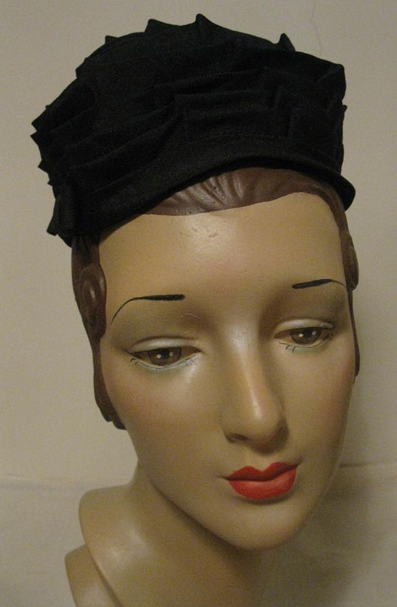 Puede incluir: Un sombrero de tela negro con un dise&ntilde;o plisado. El sombrero se lleva en un maniqu&iacute; con una expresi&oacute;n neutral y l&aacute;piz labial rojo.