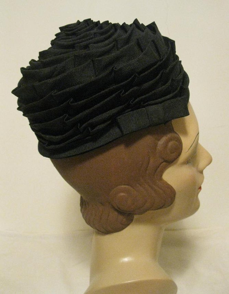 Puede incluir: Un sombrero de tela negro con un dise&ntilde;o de volantes. El sombrero se lleva en la cabeza de un maniqu&iacute;.