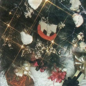 Peut inclure: Un tissu matelassé blanc avec un ornement en forme de cœur en tissu brun et blanc. L'ornement est décoré d'un ruban blanc. L'arrière-plan est un sapin de Noël vert foncé avec des ornements blancs et un traîneau rouge.