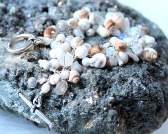 May include: A white and brown seashell bracelet with silver clasps and beads. The bracelet is on a gray rock.