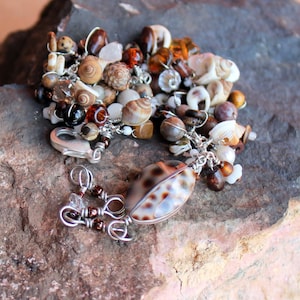 May include: A silver chain necklace with a variety of brown, white, and tan seashells, beads, and charms. The necklace has a lobster claw clasp.