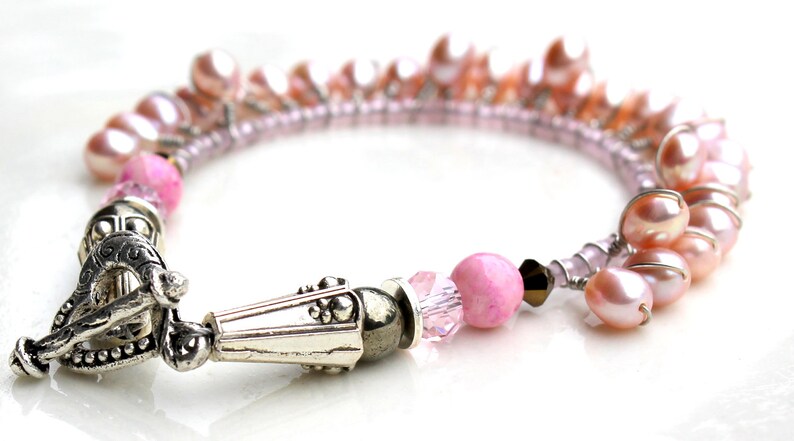Puede incluir: Una pulsera de perlas rosas con un cierre plateado. La pulsera presenta una mezcla de perlas rosas, cuentas rosas y cristales transparentes. El cierre tiene un dise&ntilde;o ornamentado.