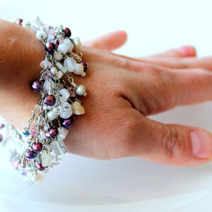 May include: A delicate bracelet with a mix of white and purple beads, shells, and crystals. The bracelet is made with silver wire and has a dangling design.