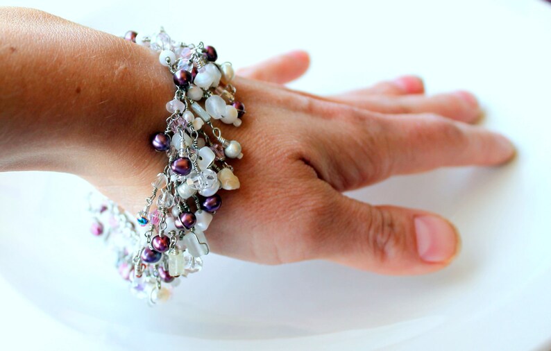 May include: A delicate bracelet with a mix of white and purple beads, shells, and crystals. The bracelet is made with silver wire and has a dangling design.