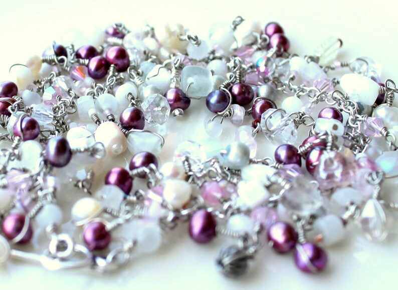 May include: A silver chain necklace with a variety of beads, including white, pink, and purple. The beads are made of different materials, including glass, crystal, and metal.