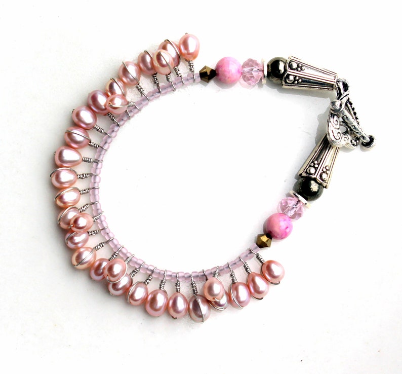 Puede incluir: Una pulsera artesanal con perlas rosas de agua dulce, peque&ntilde;as cuentas rosas y detalles decorativos de metal plateado. La pulsera tiene un dise&ntilde;o &uacute;nico con cierre de broche, sobre un fondo blanco.