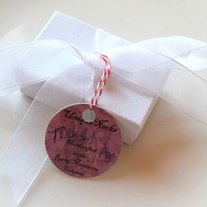 Puede incluir: Una peque&ntilde;a caja de regalo blanca atada con una cinta blanca y un cord&oacute;n rojo y blanco. Una etiqueta redonda con el texto "Unique Necks" y "Thank you! Handcrafted in the Rocky Mountains Colorado" est&aacute; adjunta.