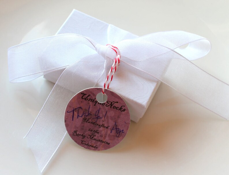Puede incluir: Una peque&ntilde;a caja de regalo blanca atada con una cinta blanca y un cord&oacute;n rojo y blanco. Una etiqueta redonda con el texto "Unique Necks" y "Thank you! Handcrafted in the Rocky Mountains Colorado" est&aacute; adjunta.