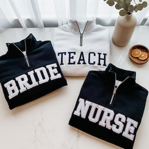 Op de afbeelding: Drie sweatshirts met een kwart ritssluiting in zwart en wit. De zwarte sweatshirts hebben "BRIDE" en "NURSE" in witte blokletters. Het witte sweatshirt heeft "TEACH" in marineblauwe blokletters. Allemaal hebben ze zilveren ritsen.