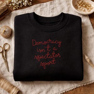 Może przedstawiać: Czarna bluza z czerwonym napisem "Democracy isn't a spectator sport". Bluza z okrągłym dekoltem jest prezentowana na neutralnej lnianej powierzchni, z akcesoriami do szycia i elementami dekoracyjnymi w tle.