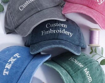 May include: A collection of custom embroidered baseball caps in various colors, including gray, blue, pink, and green. Each cap features white text, with phrases like "Custom Text," "Custom Embroidery," and "House Mom." Spools of thread are also visible.