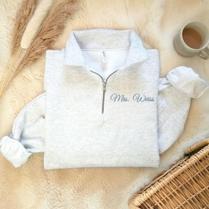 Puede incluir: Una sudadera gris claro con cuello con cremallera y el nombre "Mrs. Weiss" bordado en letra cursiva. Las mangas están dobladas y la sudadera está sobre una superficie cremosa y esponjosa. También se ven una taza de té y una cesta de mimbre.