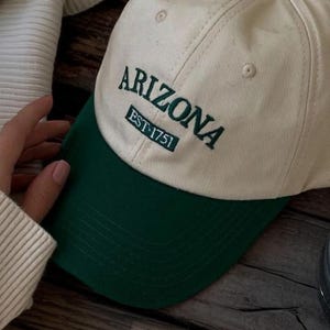 Op de afbeelding: Crèmekleurige baseballcap met een groene klep en een groene knoop bovenop. Het woord "ARIZONA" is in het groen op de voorkant geborduurd, met daaronder "EST. 1751". Een casual pet voor dagelijks gebruik.