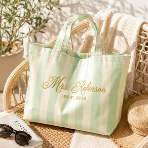 Pode incluir: Uma bolsa tote de lona com listras verticais em verde claro e branco com duas alças superiores. A frente da bolsa é bordada com o texto dourado Mrs. Robinson e o texto EST. 2026 centralizado abaixo do nome com fio dourado.