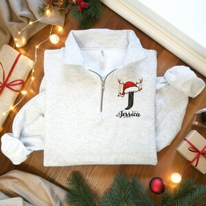 Puede incluir: Sudadera gris claro con cremallera de cuarto de cierre y diseño bordado navideño. El diseño incluye la letra negra "J" con gorro de Papá Noel y cuernos, y el nombre "Jessica" debajo. La sudadera está sobre una superficie de madera con adornos navideños.