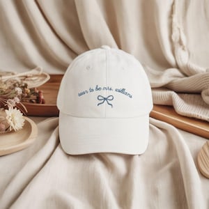 Peut inclure: Casquette de baseball blanche avec l'inscription brodée en bleu "soon to be mrs. williams", accompagnée d'un motif de nœud bleu. La casquette est en tissu doux et possède une visière incurvée.