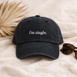 May include: A charcoal grey baseball cap with a curved brim and an adjustable strap at the back. The front of the cap features the text "i'm single." embroidered in white lowercase letters. The cap has a washed, slightly faded fabric texture.