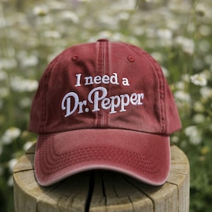 Puede incluir: Gorra de béisbol granate con la frase "I need a Dr. Pepper" bordada en blanco. La gorra está hecha de una tela suave y tiene una visera curva. La gorra está sobre un poste de madera.
