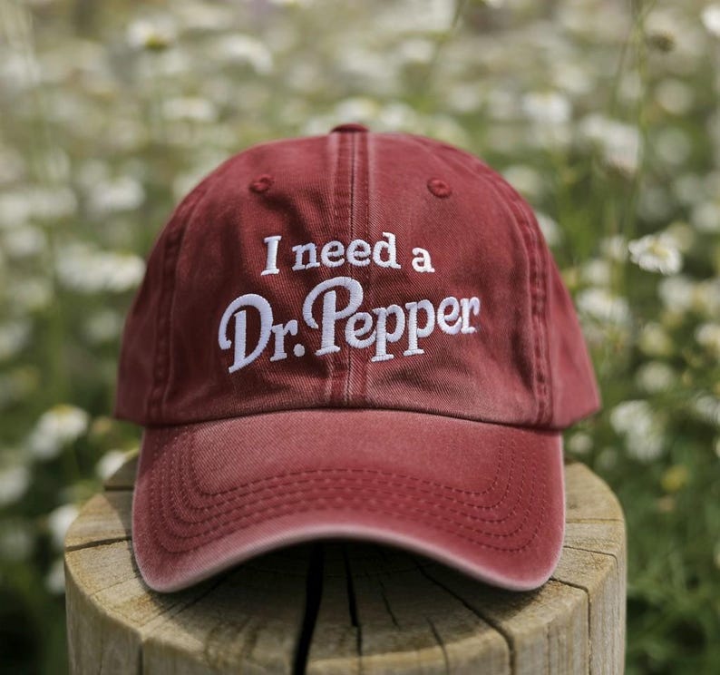 May include: A maroon baseball cap with the phrase "I need a Dr. Pepper" embroidered in white. The cap is made of a soft fabric and has a curved brim. The cap is sitting on a wooden post.