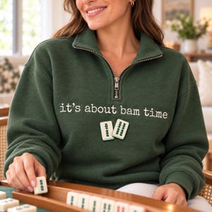 Könnte beinhalten: Ein dunkelgrüner Sweatshirt mit kurzem Reißverschluss, verziert mit dem weißen Sticktext "it's about bam time" über einer Grafik von zwei Mahjong-Steinen. Das Kleidungsstück hat einen hohen Kragen und lange Ärmel.