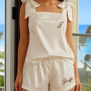 May include: Ivory-colored pajama set featuring a tank top with bow-tie straps and matching shorts. The top has a single initial embroidered on the front, and the shorts are embroidered with a name. The set is made of a soft, silky material.