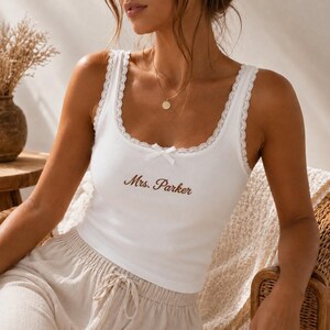 May include: A white ribbed tank top featuring a scoop neckline with delicate lace trim, a small white bow at the center, and the text "Mrs. Parker" embroidered in brown script across the chest. The garment is paired with light beige drawstring pants.