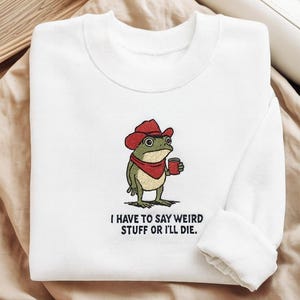 Puede incluir: Sudadera blanca con un bordado de una rana con sombrero y pañuelo rojos, sosteniendo una taza. El texto "I HAVE TO SAY WEIRD STUFF OR I'LL DIE" está debajo. La sudadera está doblada sobre una superficie beige.