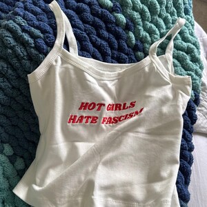 May include: White vest top with the embroidered text "HOT GIRLS HATE FASCISM" in red. The top has thin straps and a scoop neckline. It is lying on a blue and teal knitted blanket.