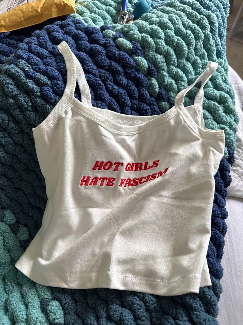 May include: White vest top with the embroidered text "HOT GIRLS HATE FASCISM" in red. The top has thin straps and a scoop neckline. It is lying on a blue and teal knitted blanket.