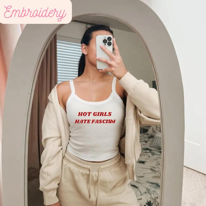 May include: White vest top with the red text "HOT GIRLS HATE FASCISM". The top has a scoop neckline and thin straps. A beige jacket and joggers are also visible in the image.