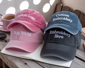 May include: Two baseball caps, one pink and one blue, are stacked on a white notebook. The pink caps have the text "Custom Logo" and "Dog MOM" embroidered in white. The blue cap has "Custom Embroidery" and "Embroidery Here" in white.