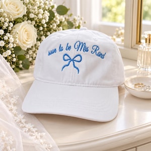 Peut inclure: Une casquette de baseball blanche avec un texte brodé en bleu indiquant soon to be Mrs Rand, accompagné d'un motif de nœud brodé en bleu centré sous le texte. La casquette présente une calotte structurée et une visière incurvée en tissu blanc.
