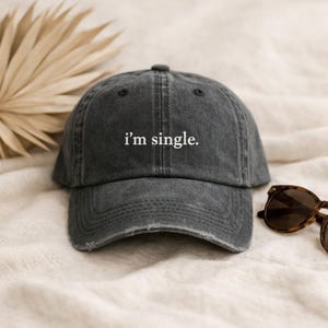 Könnte beinhalten: Eine anthrazitfarbene Baseballkappe mit dem weißen Text "i'm single.". Die Kappe hat einen Used-Look und liegt auf einer cremefarbenen Oberfläche. Sonnenbrillen liegen neben der Kappe.