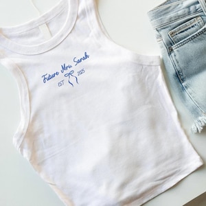 May include: White tank top with the text "Future Mrs. Sarah EST 2025" in blue script. The tank top is paired with light wash denim shorts. The top has a scoop neck and a sleeveless design.